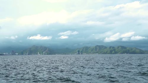 Scenic View of Wavy Sea, Bright Clouds Over Green Hills and City Buildings.