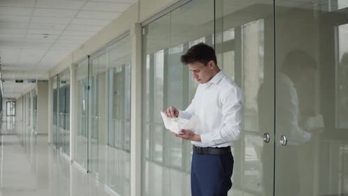 Young Office Manager Unwraps Tasty Hamburger To Eat It in Light Corridor