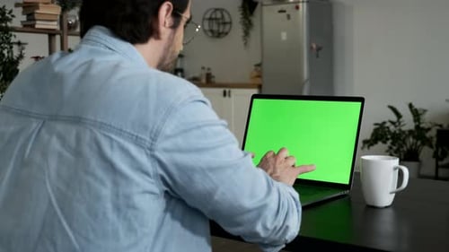 A Young Man Professional Freelancer Uses Laptop with Green Mock-up Screen While Sitting at the Desk