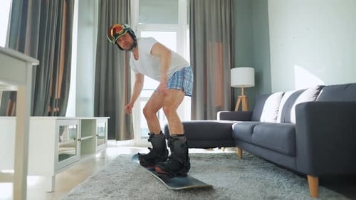 Vídeo divertido. Homem de bermuda e camiseta retrata snowboard em um tapete em um quarto aconchegante. Esperando por