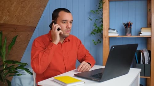 Young Man Sits in Home Office at Table with Laptop