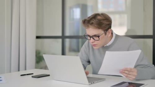 Young Man with Laptop Reacting to Loss While Reading Documents in Office