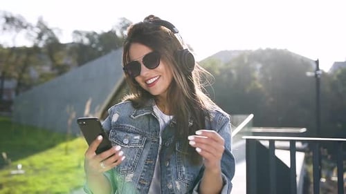 Young Woman with Glasses Using her Phone and Listening Music on Earphones on the City Background