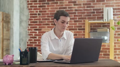 Woman with a Short Haircut and White Shirt Counts on Calculator Typing a Laptop Sitting at an Office