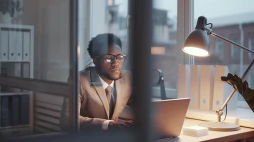 Afro-American Male Clerk Working on Laptop at Office Desk in the Evening