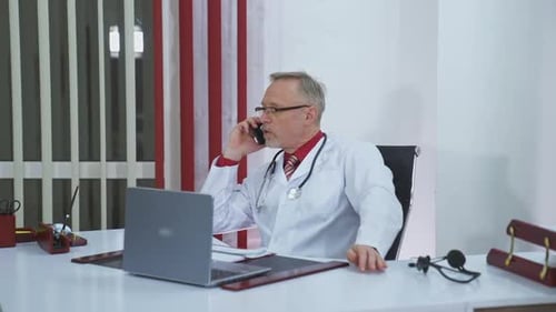 Physician talks the phone in clinic. Mature doctor in white medical coat with stethoscope sitting