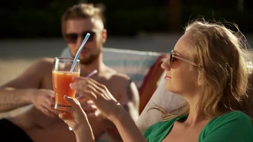 Bearded Tattoed Topless Male in Sunglasses Giving Cocktail with Straw to Blonde Female on Lounge