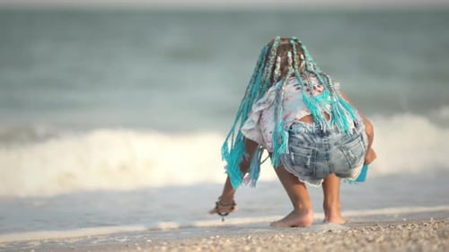A Girl with African Braids in a Summer Costume Plays on the Beach with Shells Near the Sea with