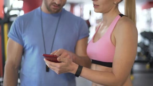 Fit Lady Showing Application Tracking Her Results to Personal Trainer in Gym