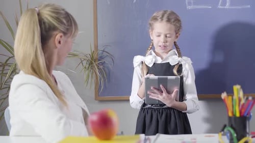 Retrato de una encantadora niña caucásica en uniforme escolar que declama a una maestra de mediana edad sentada