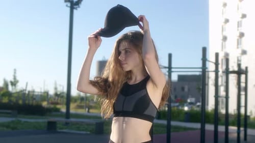 Pretty Young Woman in Black Tank Top with Wavy Hair Put on Black Cap on Street and Looks at Camera