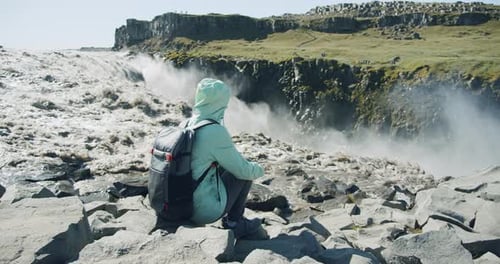 Wanderin mit Rucksack sitzt am Cliff Edge und genießt den Detifoss-Wasserfall in Island
