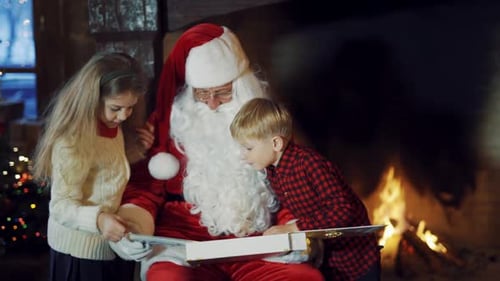 Los niños miran el álbum en manos de Papá Noel cerca de la chimenea.