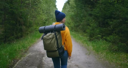 Back View of a Woman with a Backpack in a Yellow Sweater and Hat Walking Along a Road in the Woods