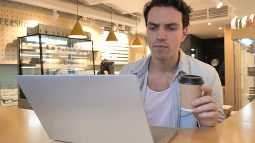 Young Man Drinking Coffee and Working on Laptop in Cafe