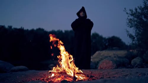 Hooded Figure Standing Near a Campfire at Night