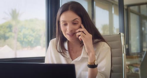 A businesswoman talking on the phone, smiling, while working on a laptop at the office