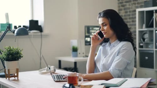 Beautiful Female Employee Chatting on Mobile Phone in Office and Using Laptop