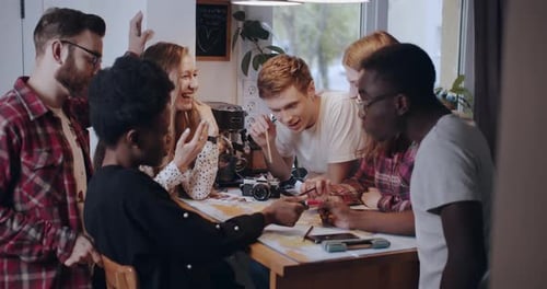 Happy Young Multiethnic Student Friends Looking at Big Map with Pencils at Kitchen Table, Planning
