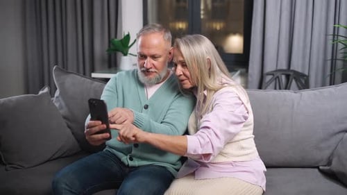 Elderly Couple View Photos on a Smartphone Sitting on the Sofa in Living Room Positive Emotions