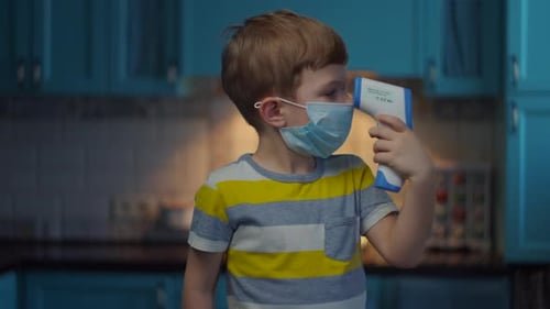 Kind mit medizinischer Gesichtsmaske misst die Temperatur mit einem Infrarotthermometer