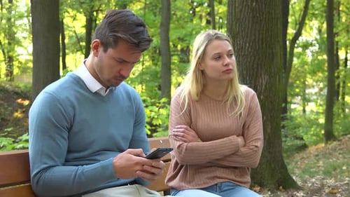 Ein Mann und eine Frau sitzen in einem Park, der Mann ist von seinem Smartphone absorbiert, die Frau ist wütend auf ihn