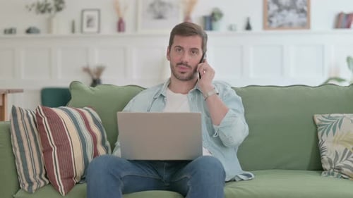 Man Working on Laptop, Talking on Phone at Home