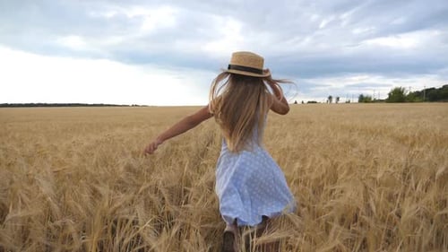 Cute Child with Long Blonde Hair Running Through Wheat Field. Little Kid in Straw Hat Jogging Over
