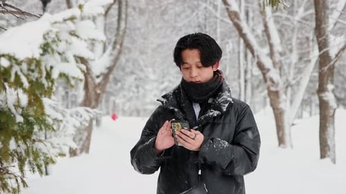 Young Asian Man Using His Phone in Winter Forest and Walks Away to Meet His Friend