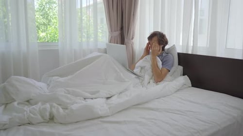 Ein trauriger junger Mann in einem Bett spricht über Videokonferenzen mit jemandem über ein Notizbuch. Er ist müde