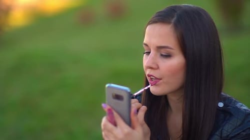 Attractive Girl in the Park Paints Her Lips, Looks at the Reflection in the Phone. Getting Ready for