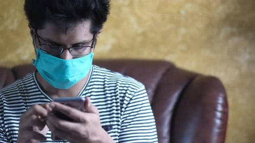 Young Man with Face Mask Using Smart Phone at Home