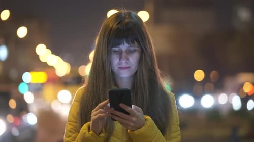 Young pretty woman using her mobile phone standing on city street at night outdoors.
