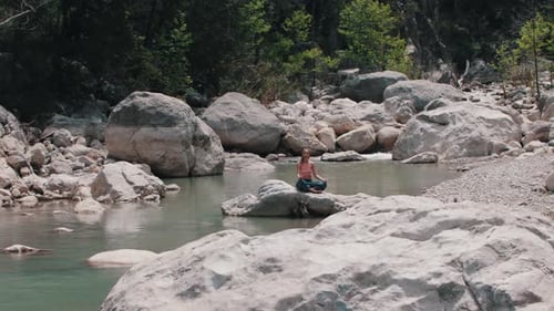 A Woman in Wide Light Pants Meditating on the Rock in the Middle of the Stream