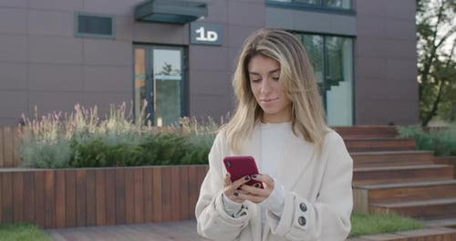 Female Attractive Person Using Smartphone While Standing at City Street. Stylish Young Woman Typing