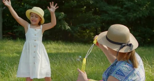 Funny Mom and Little Daughter Blow Soap Bubbles Sitting on the Lawn in the Park