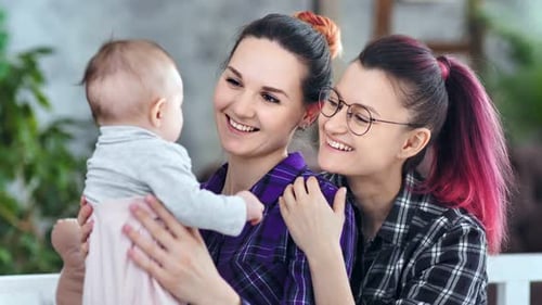 Two Women Hold Infant, Smiling Indoors