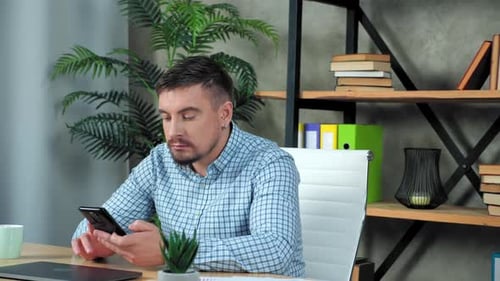 Businessman in shirt sitting on chair at desk in home office uses smartphone