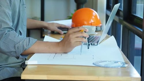 Architect is sitting in front of computer and holding divider and arrange object on desk.