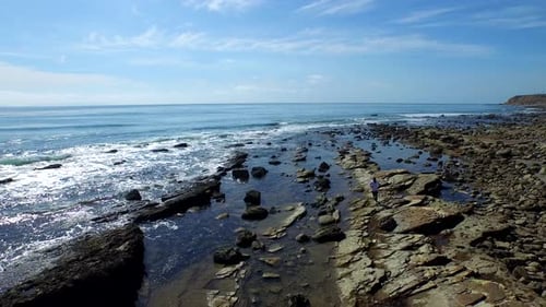Aerial birds eye view shot of a young man running on a rocky ocean beach shoreline.