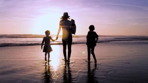 A mother holds hands and walks with her three kids at the beach in slow motion during a beautiful su