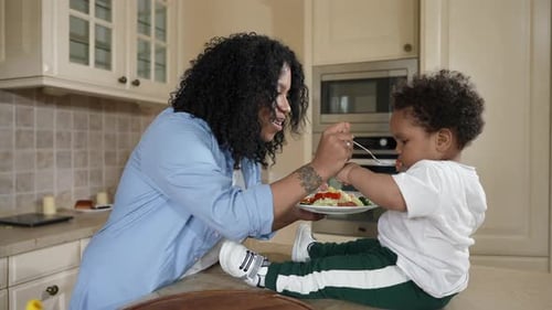 Side View Portrait Smiling African American Mother Feeding Cute Toddler Son with Healthful Vegan