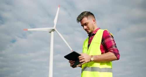 The Engineer Work Next to the Windmill Turbines with the Tablet in His Hand