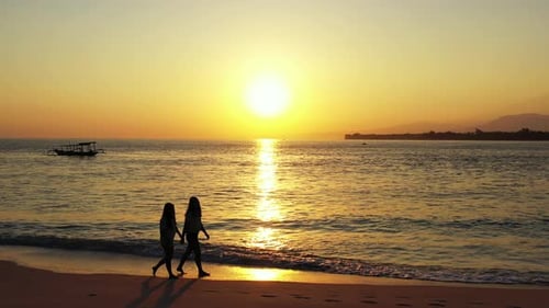 Two girls walking barefoot on sandy exotic beach washed by sea flows reflecting beautiful sunset wit