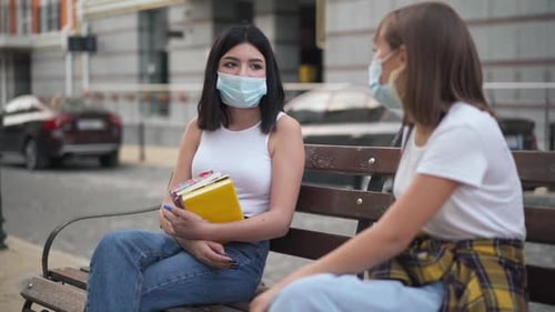 Porträt eines kaukasischen Mädchens im Teenageralter mit schwarzen Haaren in Coronavirus-Gesichtsmaske, das auf einer Bank in der Stadt sitzt