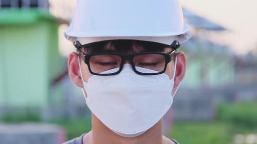 Young Asian engineer wearing a helmet and mask looks and smiles at the camera on the dam