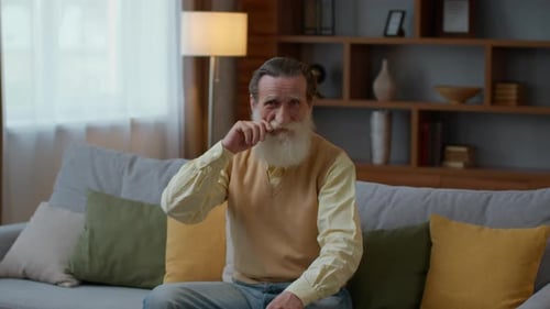 Handsome Senior Bearded Gentleman Resting on Sofa at Home Smiling to Camera and Fixing His Moustache