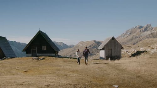 Man and Woman Couple Run By Houses Cabin in Mountain and Hold Hands Together Rbbro
