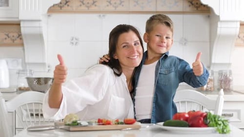 Madre e hijo guapos abrazándose y mostrando gestos con los pulgares hacia arriba en la cocina de su casa
