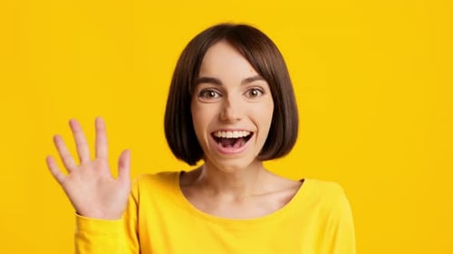 Fröhliche Millennial-Frau winkt mit der Hand und gestikuliert Hallo auf gelbem Hintergrund